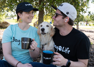 Young couple in a park smiling at each other. They are both wearing Dog Life tee shirts and hats and are holding Dog Life coffee mugs