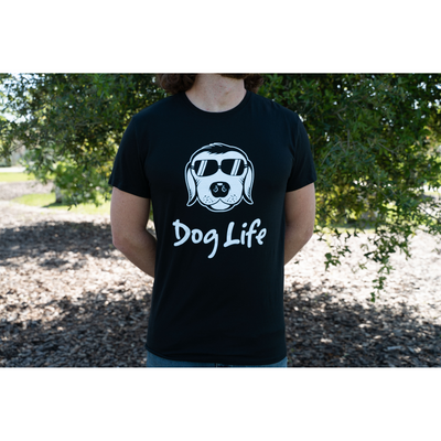 Black Dog Life tee shirt with image of a cartoon dog wearing sunglasses worn by a young man standing in front of a tree.  Torso only shown.