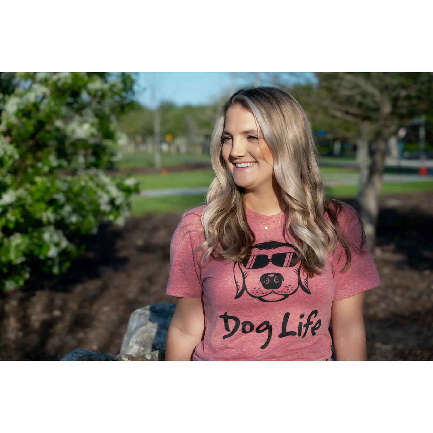  young blond Woman standing in a park and wearing Cranberry color Dog Life shirt with Classic Dog Life logo