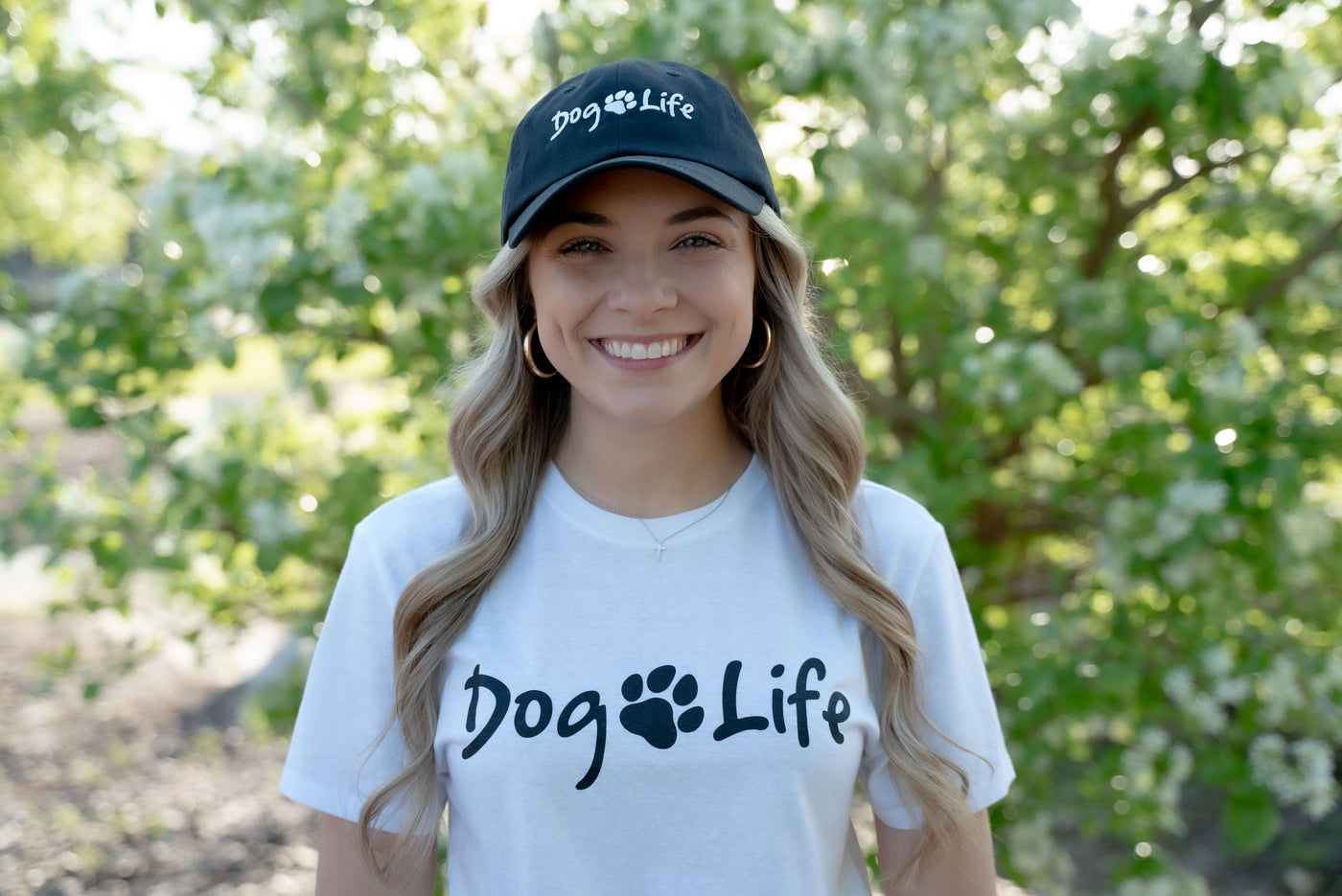 Smiling young blond woman standing outside in front of tree.  She is wearing a white tee shirt with Dog Life and a paw printed on the front.  She is also wearing a black dad hat with the Dog Life logo printed on the front.